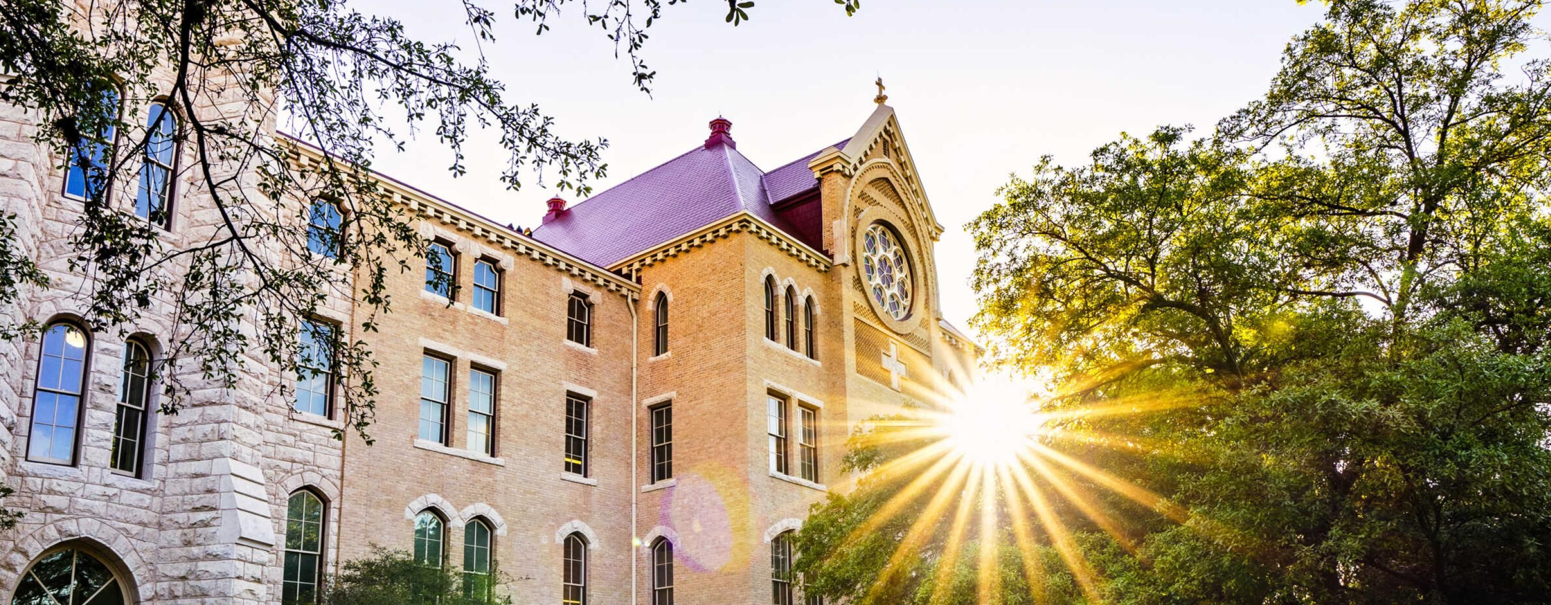 The image depicts a picturesque scene of a historic building with a clock tower, bathed in warm sunlight. The sun is partially hidden behind the building, creating a starburst effect with rays of light spreading across the scene. The building is surrounded by lush green trees, and the foreground features a spacious, empty brick-paved courtyard. The overall atmosphere is serene and inviting, likely representing a campus or significant landmark.