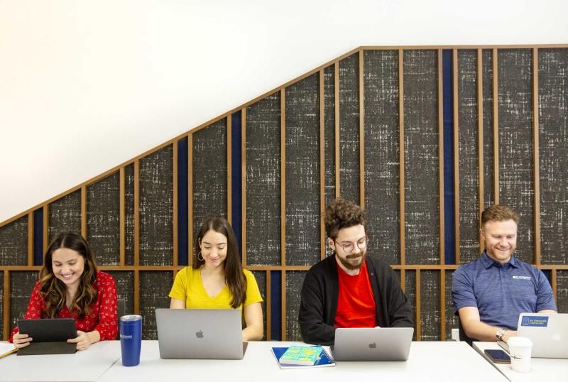 Four individuals are sitting at a table, each using different electronic devices. From left to right, the first person is using a tablet, the second and third people are using laptops, and the fourth person is using a smartphone. They are seated against a background with vertical wooden slats, behind which there appears to be mathematical equations or programming code. The context suggests an academic or work-related setting where technology is being utilized for various tasks.