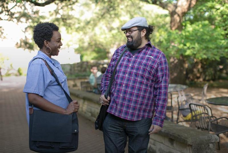 The image shows two people standing outdoors, facing each other. The person on the left is wearing a short-sleeved shirt with vertical stripes, dark pants, and is carrying a shoulder bag. The person on the right is dressed in a plaid shirt with long sleeves, jeans, and a white cap. In the background, there's a natural setting with trees and what appears to be a park area. You can see stone edging and outdoor furniture, and there are other people further in the background walking away from the camera.