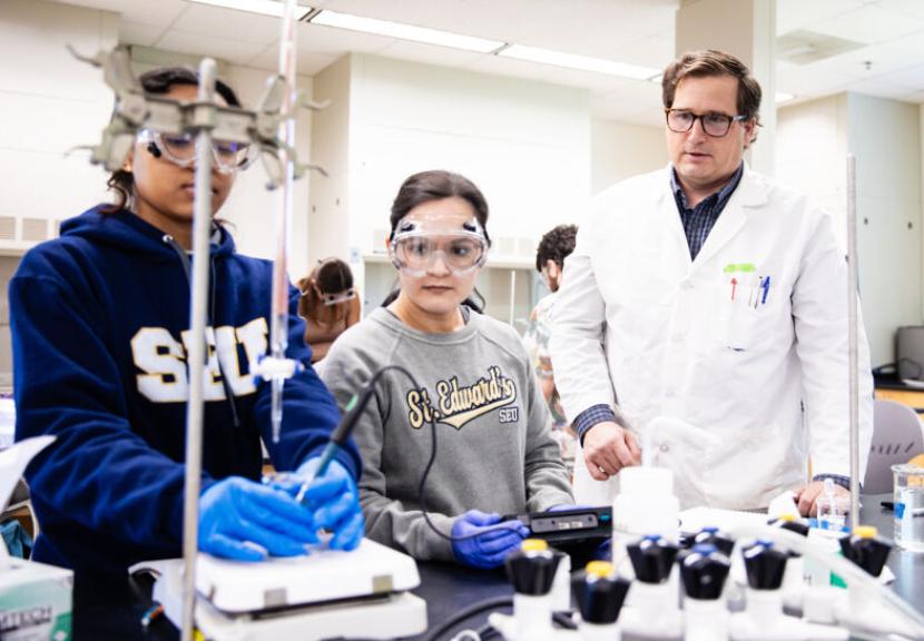 Two students wearing St. Edward's sweatshirts and a professor in a lab coat work in an analytical experiments lab.