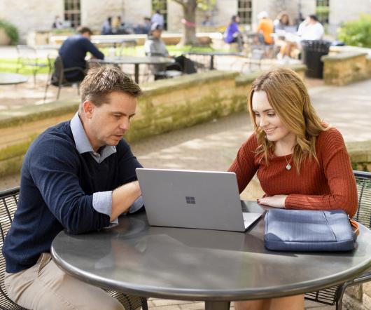 A professor and a student sit at a patio table and look at a laptop.