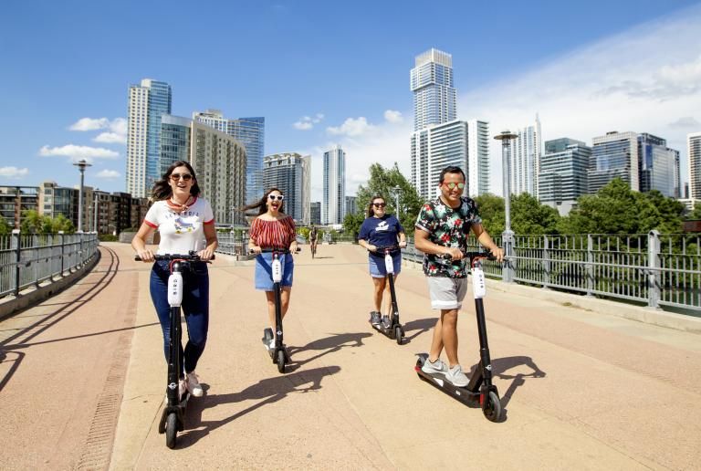 Four students ride scooters on the Pfluger Pedestrian Bridge with a view of the Austin downtown skyline behind them.