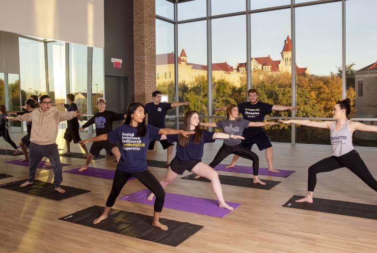 A fitness class does yoga in the fitness studio, with a view of Main Building out of the windows.