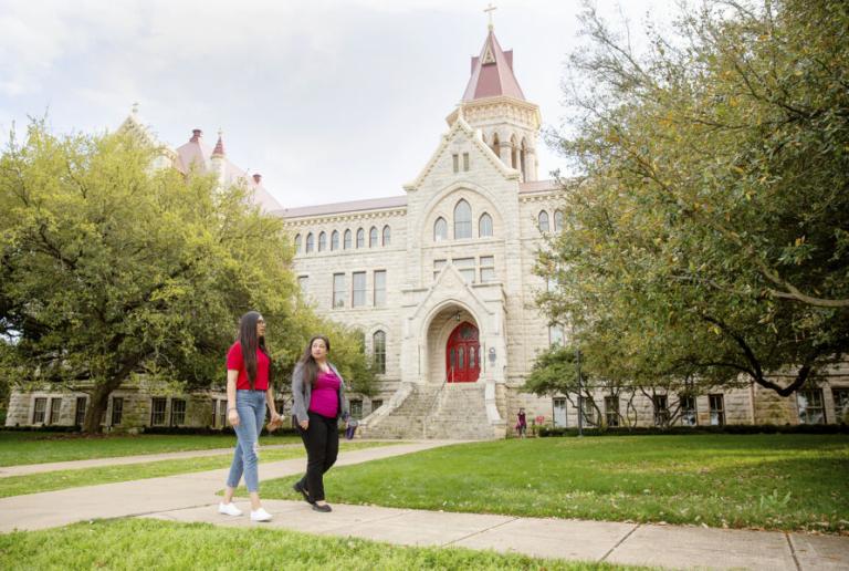 Two students walk on a path in front of Main Building and oak trees.
