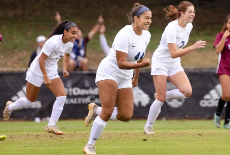 Three St. Edward's women's soccer teammates smile and run and celebrate a goal.