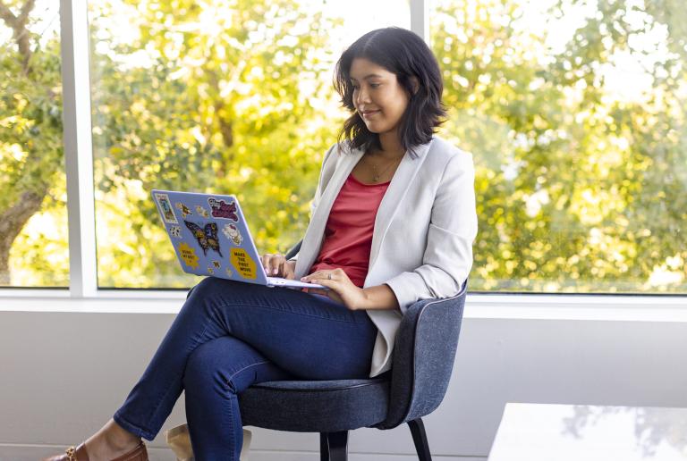 A student sits in a chair by a window with a view of trees as they work on their laptop.