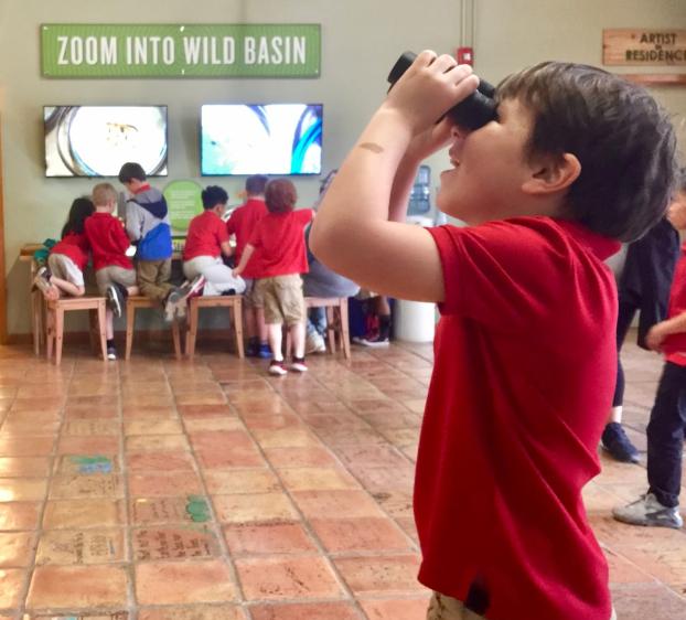 The image captures an indoor scene where a group of individuals are gathered, some seated and others standing. A child in a red shirt stands facing away from the camera, holding up a camera or phone as if taking a photograph. Two monitors display wildlife images; one shows an insect, and the other is partially visible. Above the monitors, text reads "ZOOM INTO WILD BASIN." On the wall, there's text related to an artist residency program.