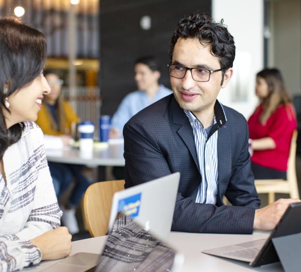 Two graduate students sit at a table in the Munday Library, chatting and studying with their laptop and tablet.