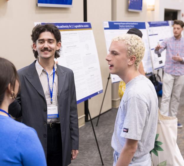 A student presenter talks with other students in front of their research poster.