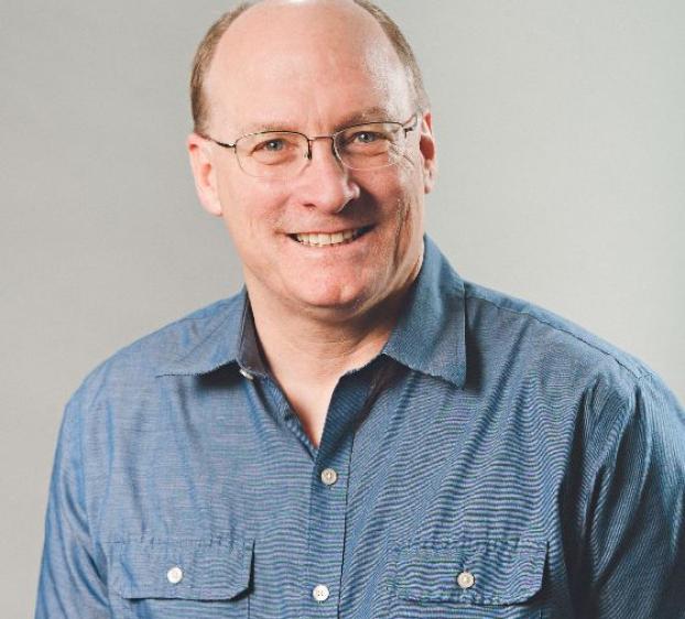 The image shows a man with a bald head and light hair on the sides, wearing glasses. He is dressed in a blue button-up shirt with front pockets. He has a friendly expression with a broad smile and is facing slightly to his right. The background is a plain light gray. He has light skin and an approachable demeanor.