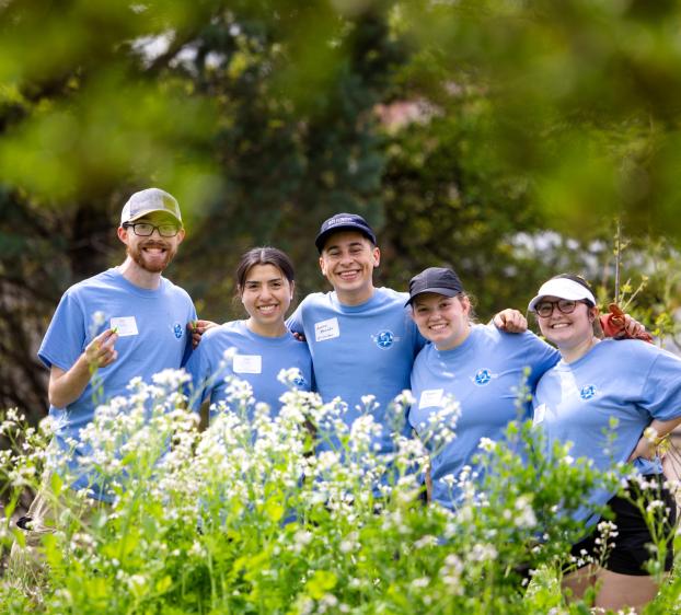 Five students wearing blue shirts stand together, framed by greenery, in the Students for Sustainability campus garden.