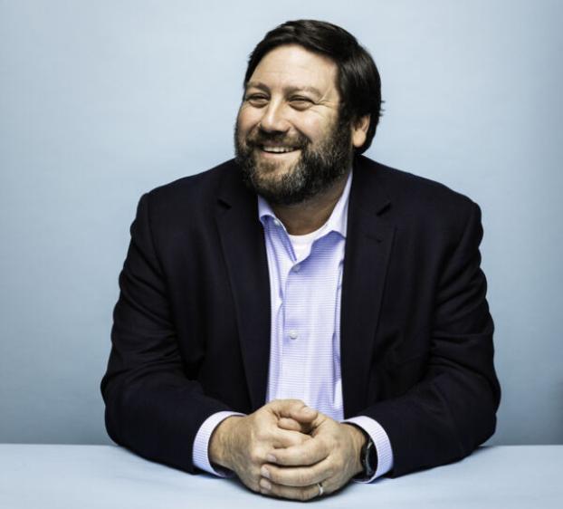 The image shows a man with a beard and short dark hair, smiling warmly and looking slightly to the side. He is seated at a table, with his hands clasped together in front of him. He is wearing a dark suit jacket over a light blue dress shirt. The background is a plain light blue, which complements his attire and keeps the focus on him. The setting appears to be a professional portrait, capturing a relaxed and approachable demeanor.