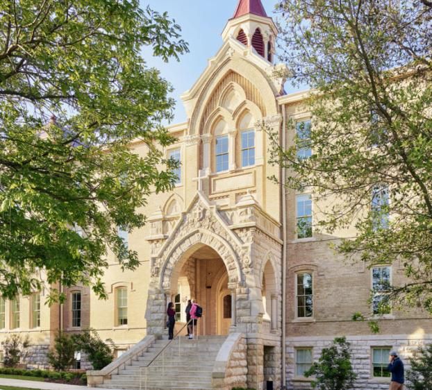 Two St. Edward's students stand outside the entrance of Holy Cross Hall as another student walks by on a path.