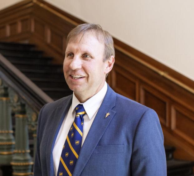 The image shows a man with short light brown hair, smiling and looking slightly to the side. He is wearing a blue suit, a white dress shirt, and a blue and yellow striped tie. The background features a staircase with ornate green and gold details and wooden paneling, suggesting the setting is an elegant, possibly historic, building. The overall impression is professional and welcoming.