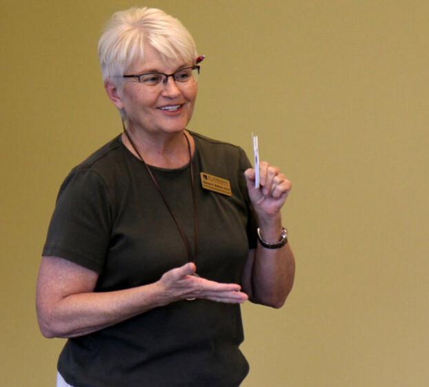 The image shows a woman with short, white hair and glasses, holding a pen or a small object while speaking. She is wearing a dark green shirt and has a name tag pinned to her chest. She appears to be in a teaching or presenting setting, gesturing with one hand and smiling. The background is plain and likely a classroom or meeting room with a neutral-colored wall.