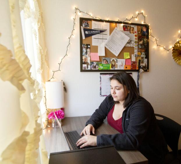 The image shows a young woman sitting at a desk in a well-decorated room. She is focused on working at her laptop. The desk is positioned next to a window with ruffled curtains, allowing natural light to brighten the space. Above the desk is a bulletin board covered with various photos, notes, and mementos, surrounded by string lights. The room has a cozy and personalized feel. A small lamp and a vase with flowers sit on the desk, enhancing the inviting atmosphere.