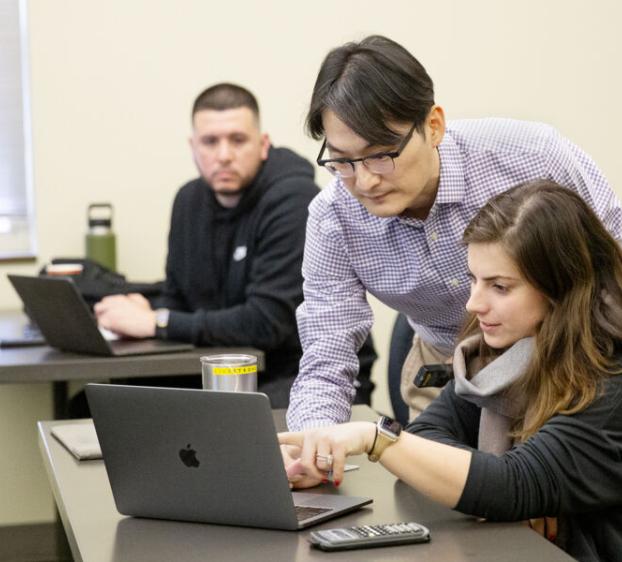 A student watches as a professor assists another student on the student's laptop in class.