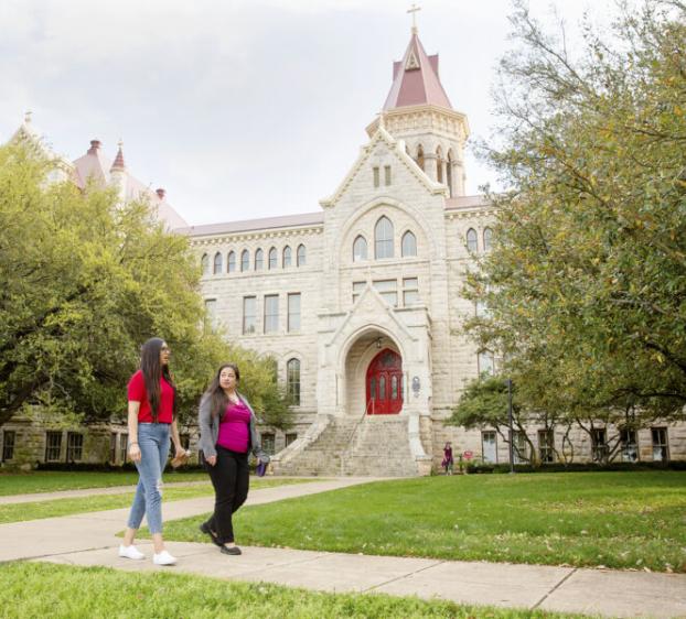 Two students walk on a path in front of Main Building and oak trees.