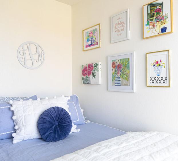 The image shows a neatly made bed with a soft blue and white color scheme. The bed is adorned with decorative pillows, including a round, pleated blue pillow in the center. Above the bed, the wall is decorated with framed art pieces and prints, featuring floral and inspirational themes. The artwork adds a colorful and cheerful touch to the minimalist, bright room. A round monogrammed piece of art is also present on the wall. The overall atmosphere is cozy and inviting, with a clean and elegant aesthetic.