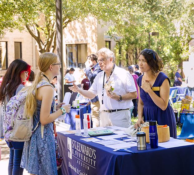 Faculty from the School of Behavioral and Social Sciences stand at a table on Ragsdale Lawn and talk to students.