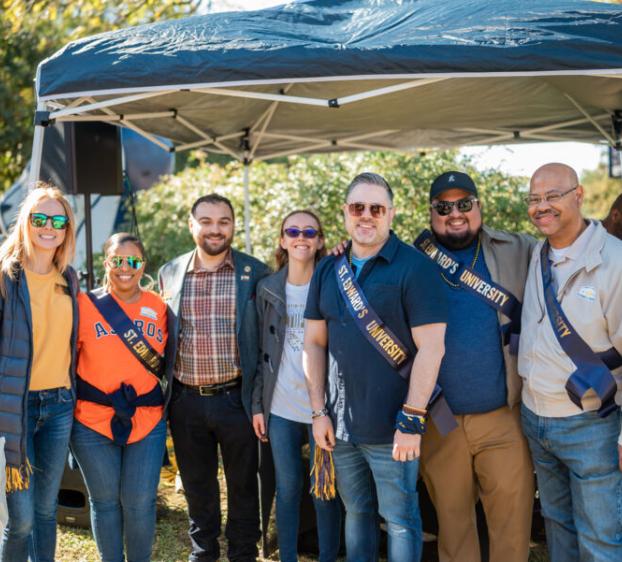 A group of people smiles for a group photo under a tent at an outdoor event, wearing sashes and crossbody bags. The setting is sunny, with trees and event booths in the background.