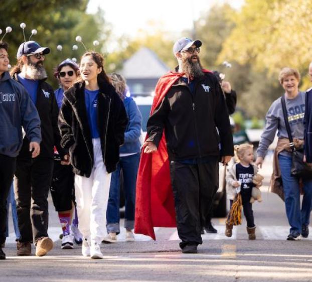 This image shows a group of people walking together in a festive setting, likely a parade or community event. They are dressed casually and appear joyful. Some wear novelty headbands with bobbing antennas. One man wears a red cape and sunglasses, adding a playful touch. A small child is holding hands with adults and looks excited. Trees and buildings are visible in the background, suggesting a suburban or park area. The atmosphere is cheerful and communal.