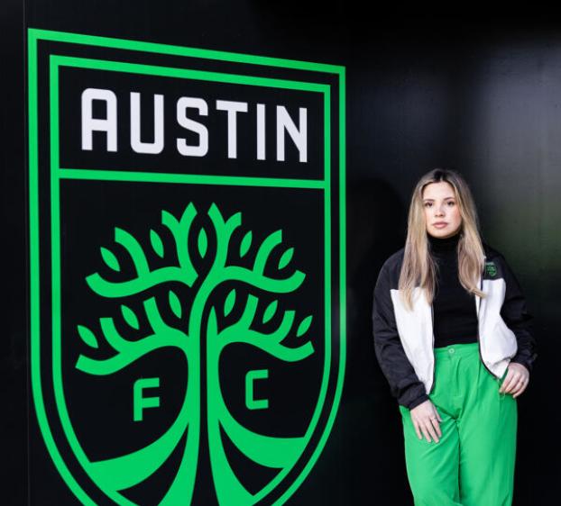 An alumna, now a staff member at Austin FC, stands by an Austin FC logo.