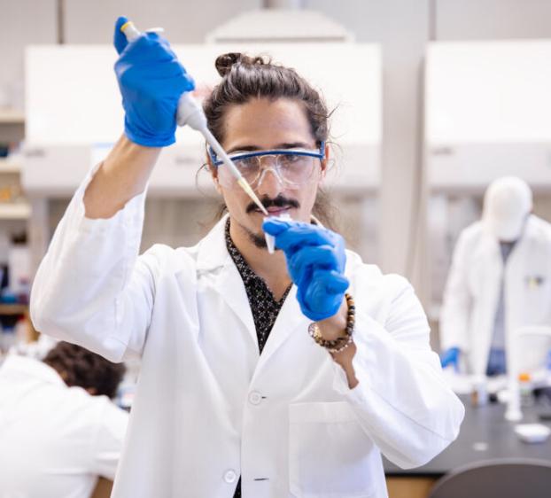 A student wearing a lab coat, protective glasses and gloves pipettes a sample in a biochemistry lab.