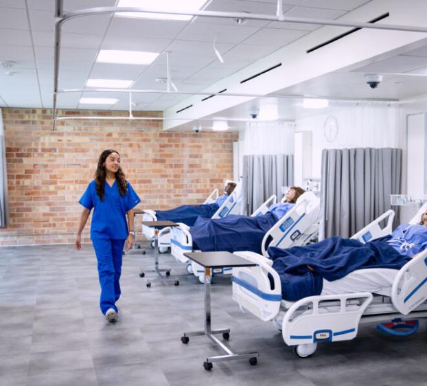 The image shows a healthcare setting, likely a hospital or a medical training facility. A nurse in blue scrubs is walking through a room with several hospital beds, each occupied by mannequins that simulate patients. The beds are equipped with medical monitoring devices, and the room has a clean, modern appearance with bright lighting and a mix of brick and white walls. This setting appears to be designed for medical training or simulations.