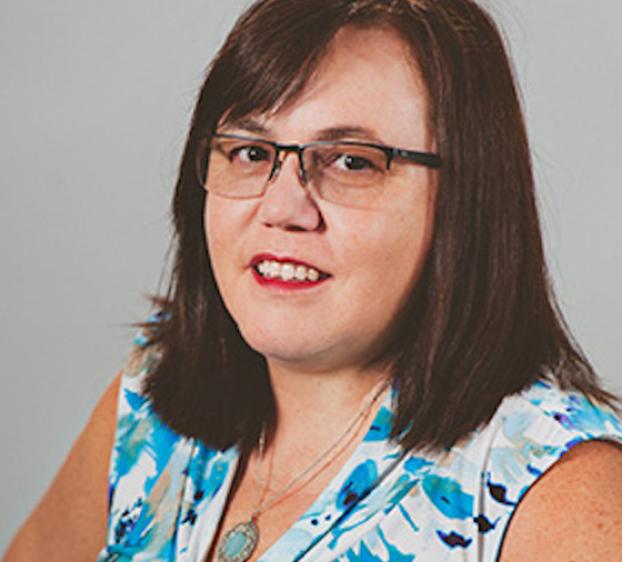 The image shows a woman with medium-length dark brown hair and wearing glasses. She is dressed in a sleeveless blouse with a blue and white floral pattern. She has a friendly expression with a slight smile and red lipstick. The background is a plain light gray. She is wearing a necklace with a round pendant.