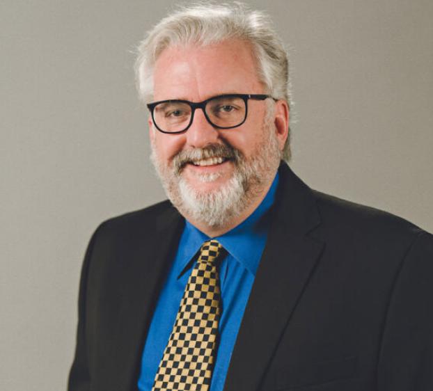 The image shows a man with gray hair and a beard, wearing glasses, a blue dress shirt, a checkered tie, and a black suit jacket. He is smiling and facing the camera against a plain background. The setting suggests it is a professional portrait, likely for business or academic purposes.