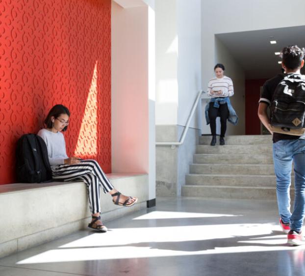 A St. Edward's student sits with their backpack on a red wall as other students walk up and down stairs in Equity Hall.