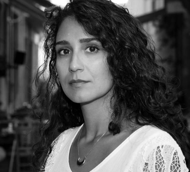 This is a black and white portrait of a woman with curly, dark hair, gazing directly at the camera with a serious expression. She is wearing a light-colored blouse with lace detailing on the sleeves and a necklace with a crescent-shaped pendant. The background appears to be an outdoor setting, slightly out of focus, with what might be trees and buildings in the distance. The overall mood is calm and introspective.