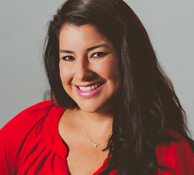 The image shows a woman with long, dark hair wearing a red blouse. She has a friendly expression with a broad smile and is facing slightly to her left. The background is a plain light gray. She has light skin and is wearing a delicate necklace.