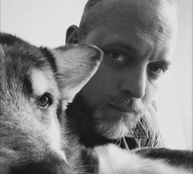 This black and white image features a close-up selfie of a man with a shaved head and beard, looking directly at the camera with a thoughtful expression. Next to him is a dog, possibly a husky or similar breed, also facing the camera. The man and the dog are positioned close together, creating a sense of intimacy and connection between them. The overall mood is warm and personal.