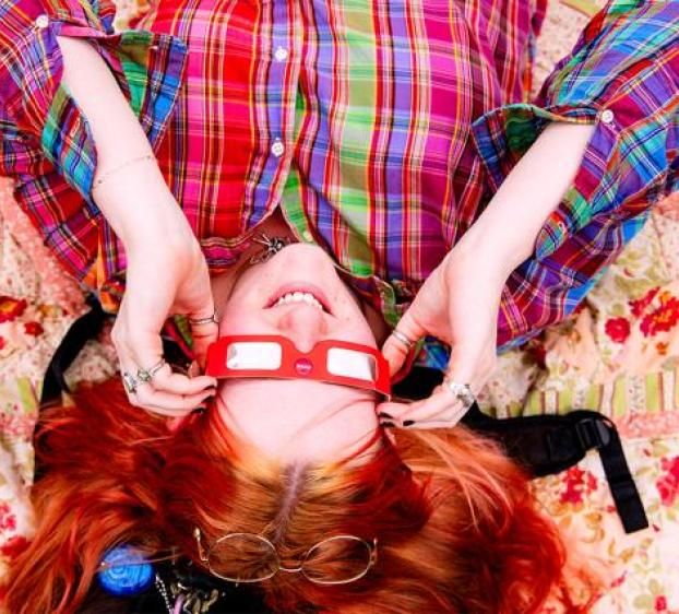 The image shows a person lying on their back on a floral quilt, looking up at the sky through special eclipse glasses. The person is smiling and appears relaxed, wearing a colorful plaid shirt that matches the vibrant, patterned quilt beneath them. The glasses, designed for safe solar viewing, suggest that the person is preparing to watch or is in the middle of watching a solar eclipse. The scene captures a joyful and carefree moment.