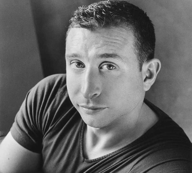 This black and white portrait features a man with short, curly hair and a clean-shaven face, looking directly at the camera with a slight, confident smile. He is wearing a simple, dark-colored T-shirt. The background is blurred and neutral, keeping the focus on the subject. The overall mood of the image is relaxed and approachable, with the man’s expression conveying a sense of warmth and openness.