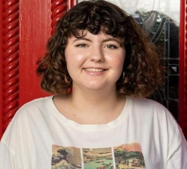 The image shows a young woman with curly, dark hair, smiling at the camera. She is wearing a white t-shirt with a colorful graphic print on the front. The background features a red door with ornate details and glass panels, giving a vibrant and visually interesting setting. The overall mood of the image is friendly and welcoming.