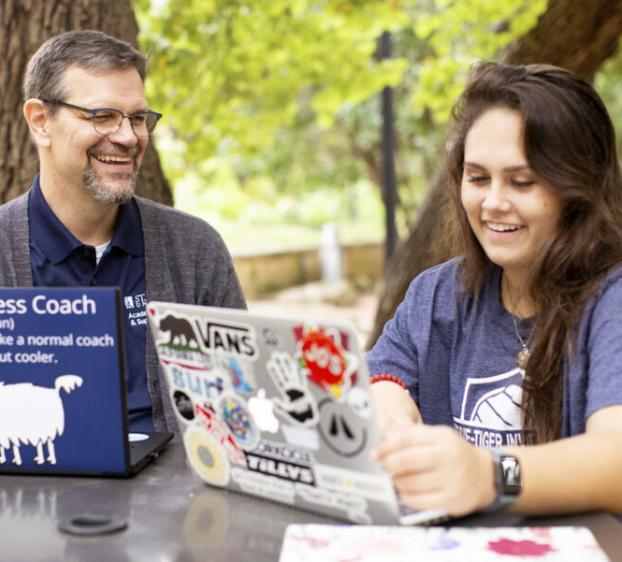 A success coach and a student, both smiling, on their laptops at a table outside