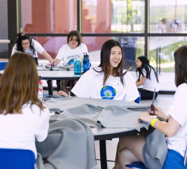 A group of students sits around tables, laughing and working together on a project. They are cutting gray fabric and are wearing white "The Big Event" t-shirts.