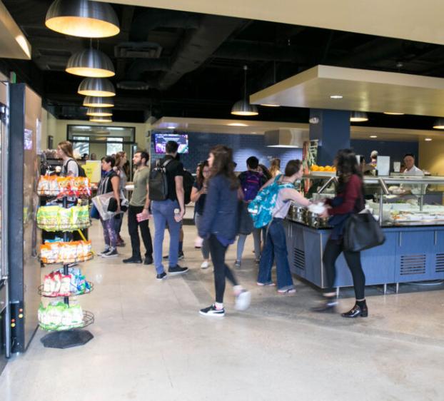 The image shows a busy dining area, likely in a cafeteria. People are lined up at a serving station where food is being prepared and served. On the left, there is a refrigerated section stocked with beverages and snacks, and a display with various packaged food items. The atmosphere is lively, with several students or patrons, some carrying backpacks, moving through the space, getting food, and interacting with staff.