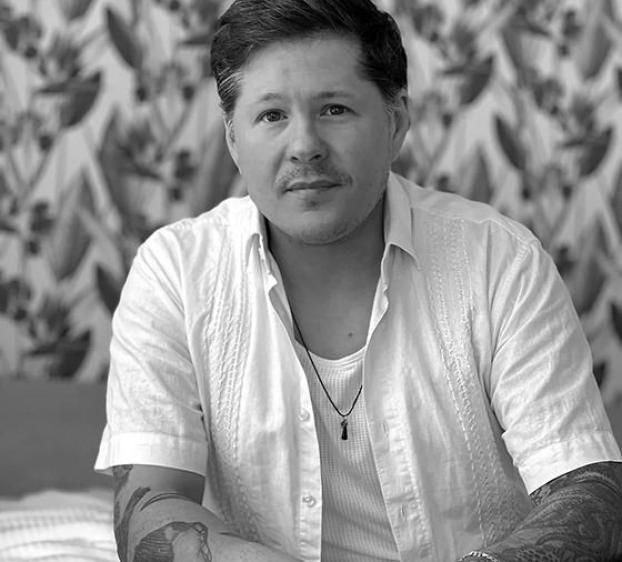 The image shows a man sitting indoors, wearing a short-sleeve white shirt over a white tank top. He has short dark hair and a mustache, with visible tattoos on his forearms. A necklace with a small pendant rests on his chest. The background features a decorative, leafy wallpaper. The man appears calm, looking directly at the camera. The image is in black and white.