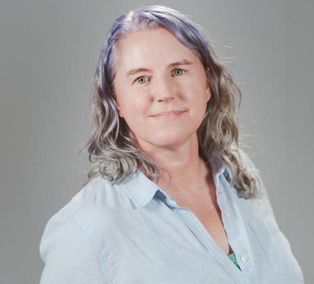 A professional headshot of a woman with shoulder-length, wavy hair that is silver with purple highlights. She is wearing a light blue button-down shirt over a green top and has a soft, neutral expression. The background is a solid, light gray.