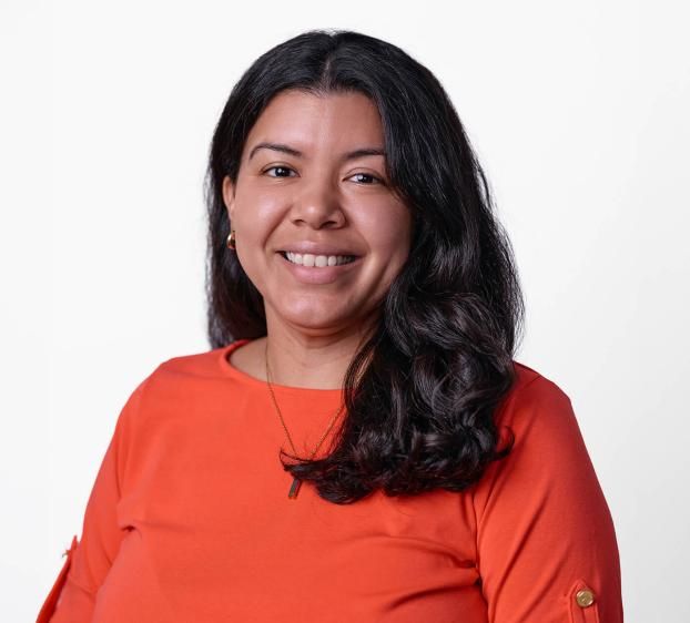 A professional studio headshot of a smiling woman with long, wavy dark hair. She is wearing a vibrant orange short-sleeved blouse and a delicate gold necklace. She stands against a clean, solid light grey background, looking directly at the camera with a friendly expression.