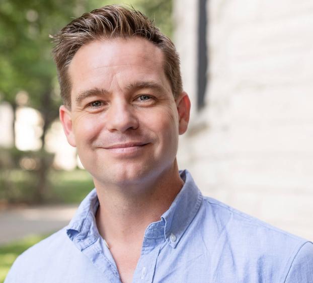 A headshot of a smiling man with short, styled brown hair and fair skin. He is wearing a light blue button-down shirt. The background is softly blurred, showing a white limestone wall on the right and green trees with sunlight on the left.