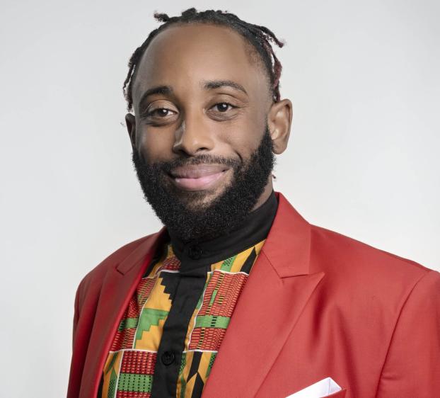 A headshot of a smiling man with a well-groomed beard and short, dark locs with reddish-brown tips. He is wearing a vibrant red blazer over a colorful patterned shirt featuring traditional African geometric designs in yellow, green, and black. He has a confident, friendly expression and is posed against a plain, light grey background.