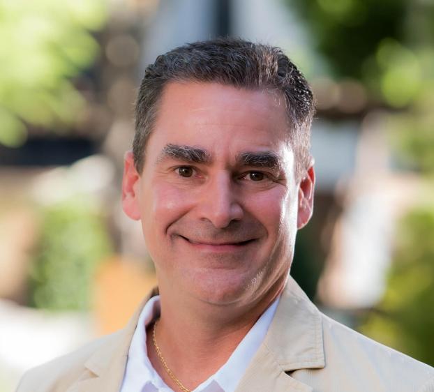 A close-up headshot of a man with short, dark, salt-and-pepper hair and brown eyes. He is smiling warmly at the camera. He wears a tan blazer over a white collared shirt and a thin gold chain necklace. The background is a soft, out-of-focus outdoor setting with green foliage and natural sunlight.