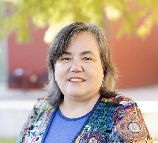 Gemini said A headshot of a smiling woman with shoulder-length, salt-and-pepper hair. She is wearing a blue top under a vibrant, multi-colored jacket featuring intricate circular and floral patterns. She has fair skin and is wearing small purple earrings. The background is softly blurred, showing a reddish-brown building and green tree foliage in natural daylight.
