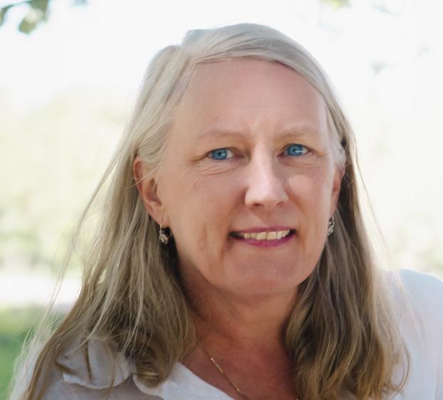Close-up portrait of a woman with long, fair hair and blue eyes, smiling gently. She is wearing a white top, small hoop earrings, and a thin gold necklace. The background is a soft-focus, bright outdoor setting with greenery and natural light filtering through trees.