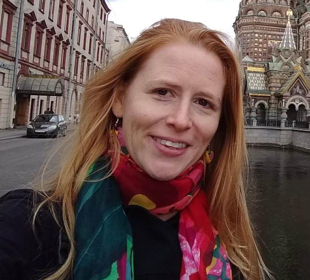 A woman with long strawberry-blonde hair and a vibrant, multicolored floral scarf smiles in a selfie. She stands on a bridge over a canal in St. Petersburg, Russia. To her left is a classic European street with a dark car, and to her right is the ornate, colorful facade of the Church of the Savior on Spilled Blood under an overcast sky.